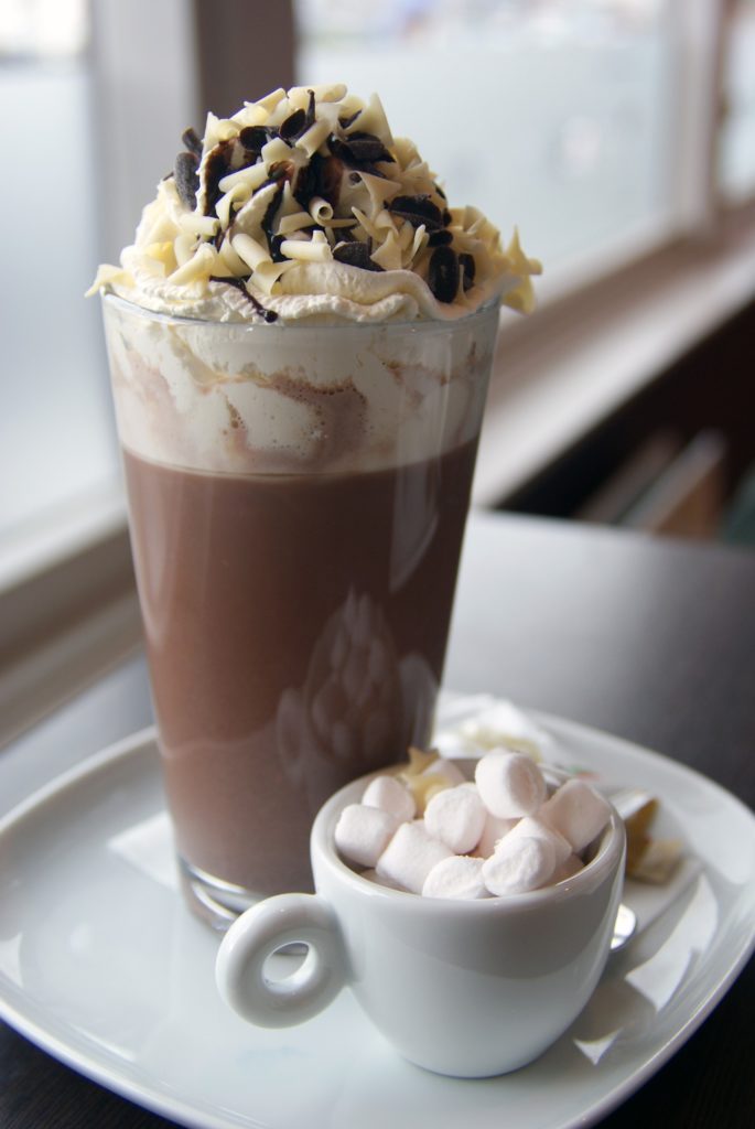 Hot chocolate in tall glass with cream and sprinkles with a separate pot of mini marshmallows on the side