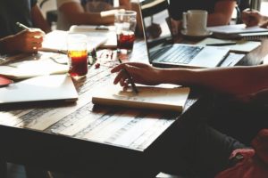 Meeting at a desk, with notebooks, drinks and laptops (you can't see their faces, just there arms writing in their books)